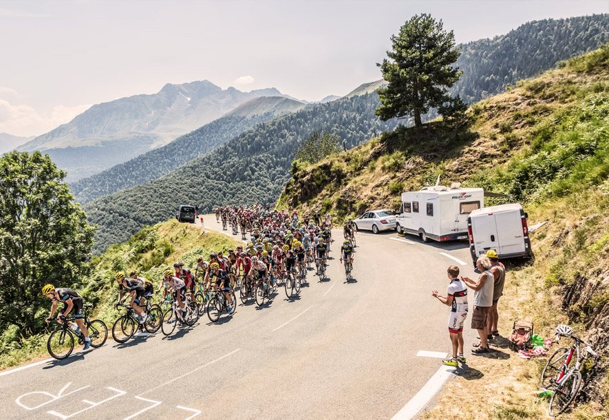 The Tour de France riders cycling on a road with dense trees and mountains behind them