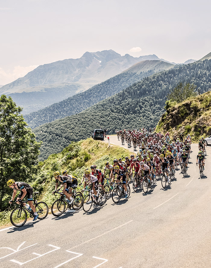 The Tour de France riders cycling on a road with dense trees and mountains behind them
